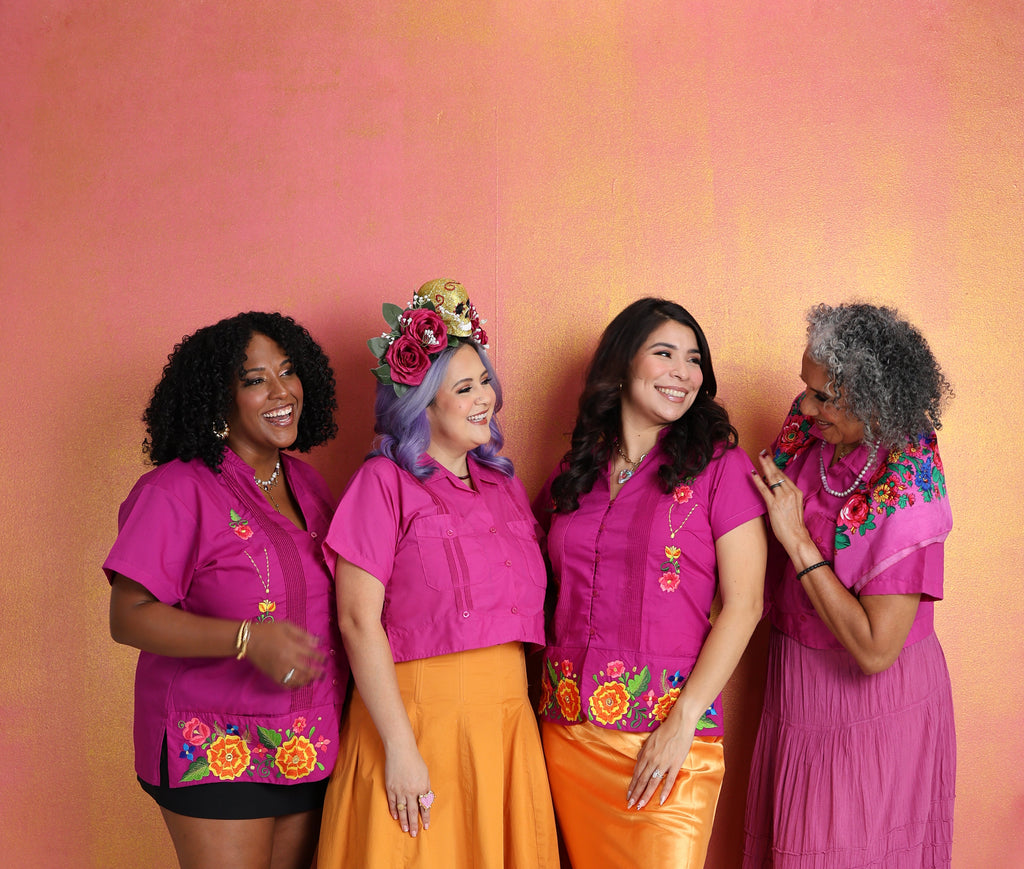 Four women in matching pink tops with colorful embroidery and orange skirts against a pink background